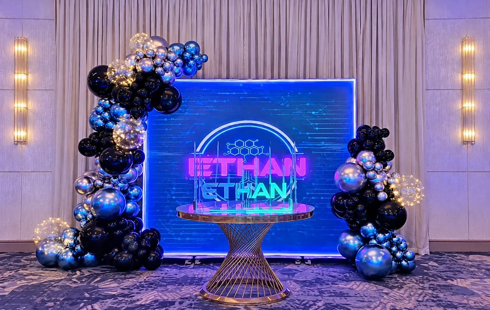 Boy’s Bar Mitzvah decor in Los Angeles with custom illuminated blue backdrop, black, chrome blue and metallic silver organic balloon garlands, clear LED light balloons, personalized neon-style display and gold table.